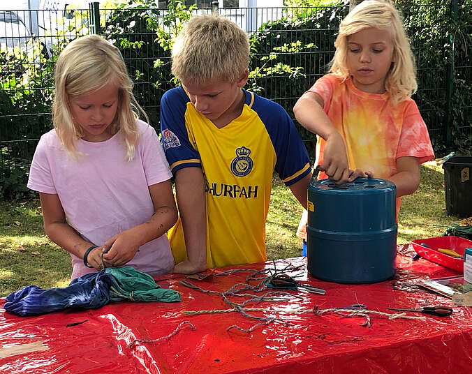 Zwei Mädchen und ein Junge stehen am Tischm it roter Tischdecke, vor ihnen ein blauen T-Shirt das sie mit Batiktechnik bearbeiten.